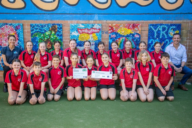 A group of 19 students kneel in front of a colourful wall while showing two fake cheques. A female adult sits on one side and a male adult sits on the other.