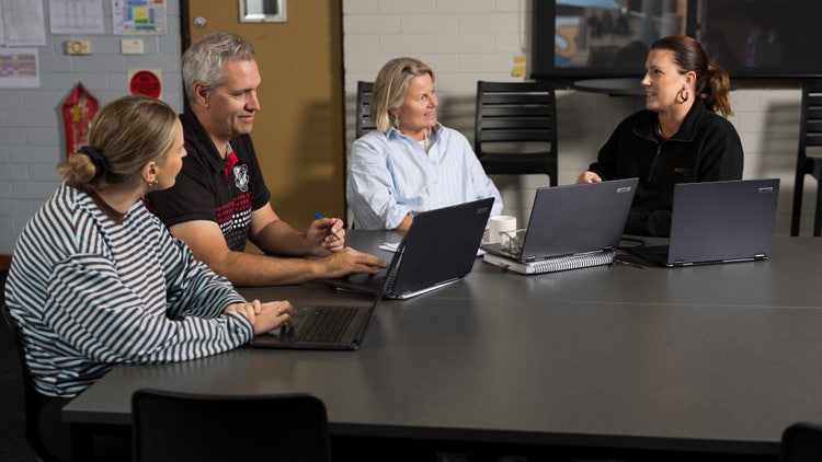 One male teachers and three female teachers sit at a desk with their laptops. One teacher appears to be speaking to the group.