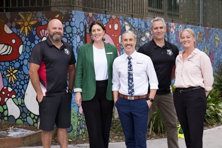 All five leaders of the Dudley Public School smile and look towards the camera. They are standing in front of a small garden with a brightly coloured mural painted behind them depicting patterns with mushrooms, birds and flowers.