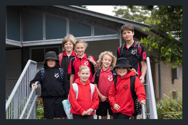 Seven students standing on the front steps to the school. The school hall is in the background.