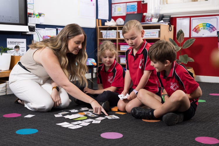 A female teacher crouches on a classroom floor and explains a concepts to three students. She is pointing to some cards spead out on the floor.