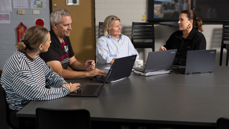 Four teachers sit at a desk with laptops. There is one male and three female teachers. The female teacher on the right appears to be directing a discussion.