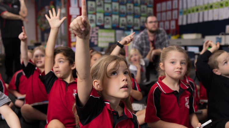 A class of kindergarten students are sitting on the floor. Several have their hands up. An SLSO sits watching the lesson from behind the students.