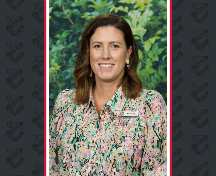 A female staff member in a floral shirt smiles for the camera with a blurred leafy background.