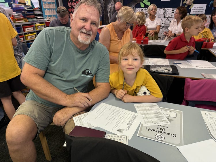 A grandfather sits at a desk with his granddaughter with worksheets in front of them.