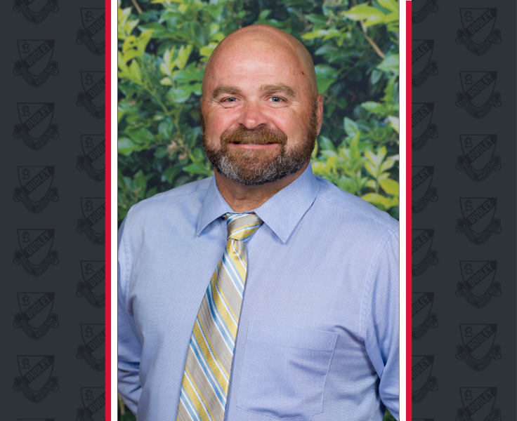 A bald male with a short full-faced beard, wearing a shirt and tie smiles looking at the camera.