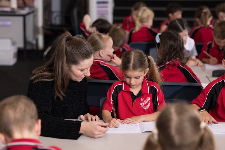 A teacher in a black top helps a young female student with her work. Other students are in the background.