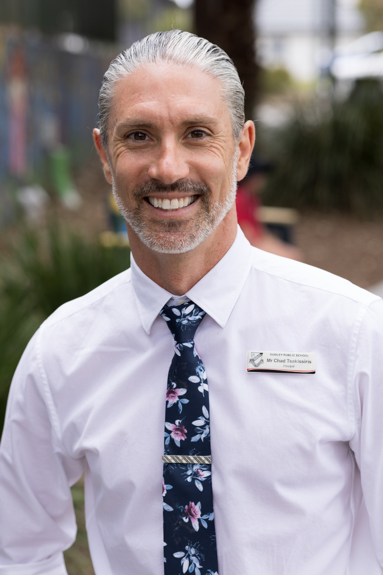 Our school Principal Mr Chad Tsakissiris smiling and looking directly into the camera.He has grey hair tied back in a bun and a short grey full-faced beard.