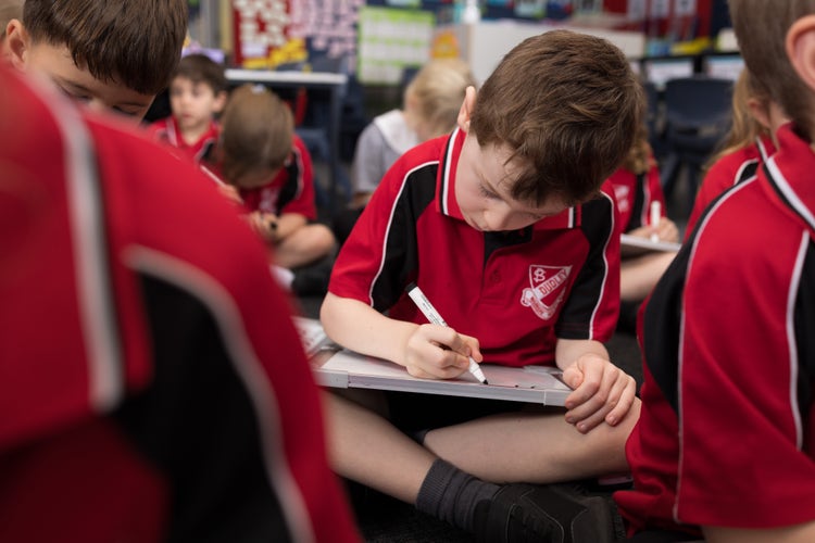 A young boy is intently writing on a whiteboard that is on his lap. Other students are in the background and blurred to the sides.