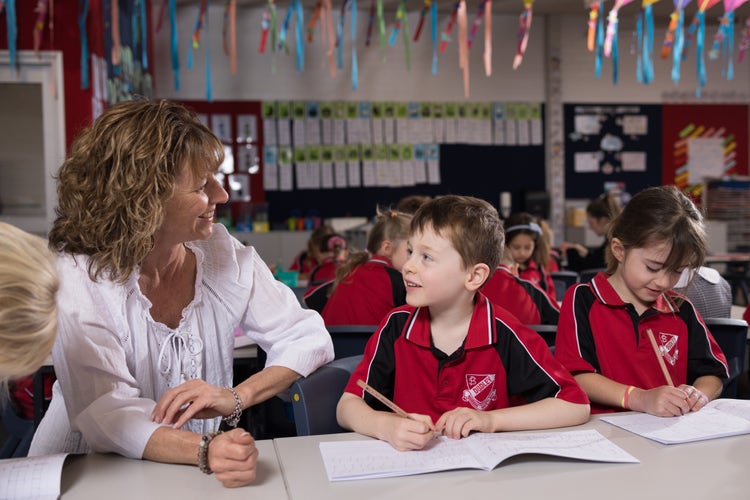 A female teacher in a white top looks at a male student and he is looking at her also. They are sitting at a desk and the boy is writing in a book.