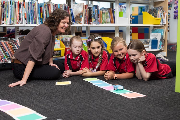 A teacher is sitting on the floor while four girls lay on the ground looking at a spherical robot and some cards laying on the carpet before them.