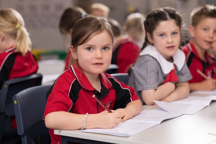 A young girl is writing in a book at her desk, looking straight at the camera. A boy and girl sit beside her slightly blurred and out of focus.