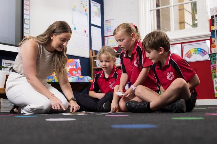 Three students watch a teacher demonstrate a maths activity on the floor.